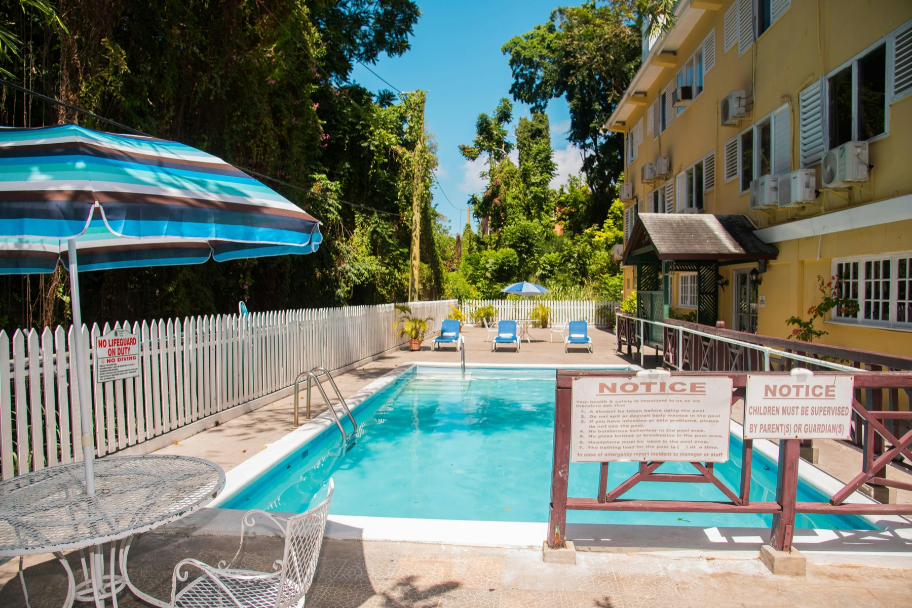 Pineapple Court's swimming pool under a striped umbrella, with the yellow hotel building beside it